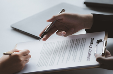 Zwei Hände, die einen Vertrag mit dem Titel "CONTRACT AGREEMENT" halten, während eine Person mit einem Stift darauf zeigt. Im Hintergrund ist ein Laptop sichtbar.